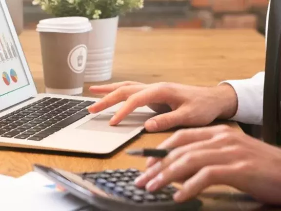 Funcionário de camisa social e gravata trabalha em um computador portátil (notebook), com uma calculadora ao lado