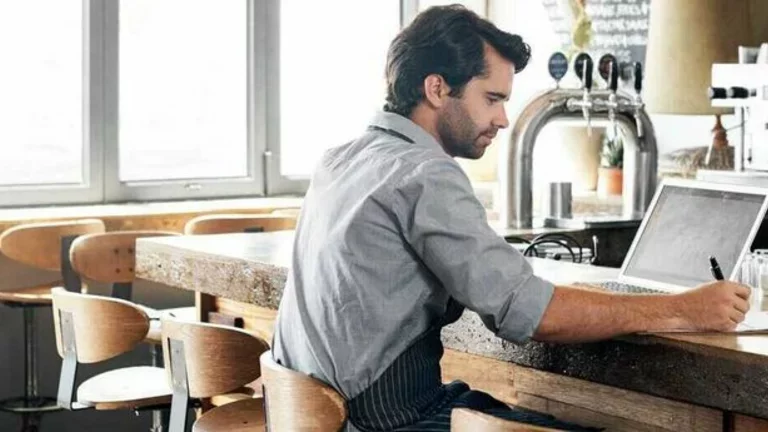 No balcão de um bar, onde há visivelmente uma chopeira e várias canecas de vidro, um homem de avental trabalha em um computador portátil (notebook) e anota com uma caneta em uma prancheta