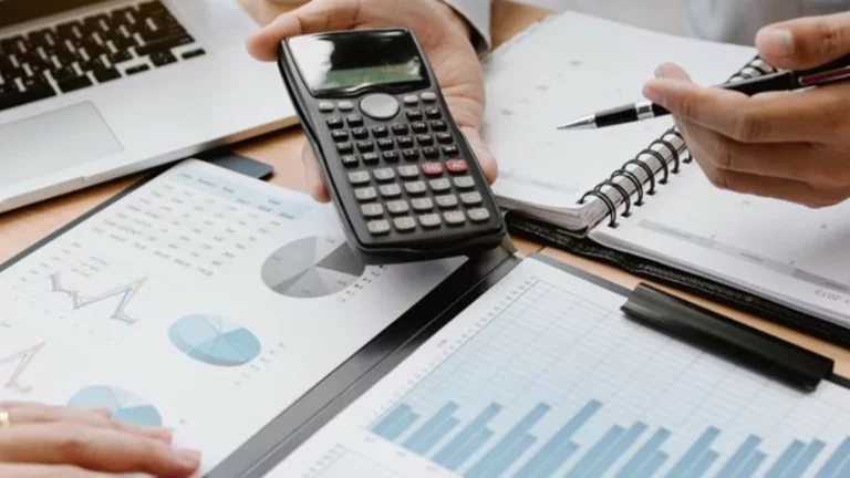 Profissionais utilizam cadernos, pranchetas com gráficos, um computador portátil (notebook) e uma calculadora para trabalhar