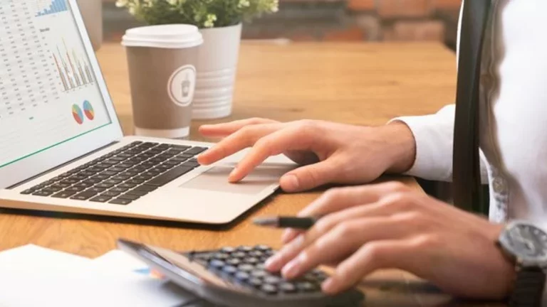 Funcionário de camisa social e gravata trabalha em um computador portátil (notebook), com uma calculadora ao lado