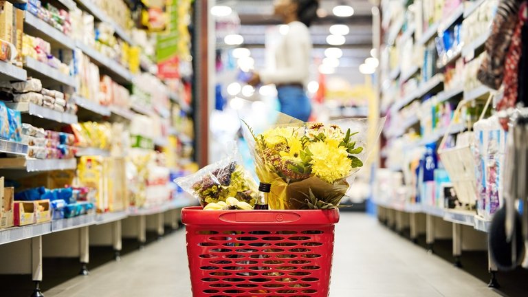 Uma cesta de compras no chão de um supermercado, enquanto uma mulher faz compras ao fundo