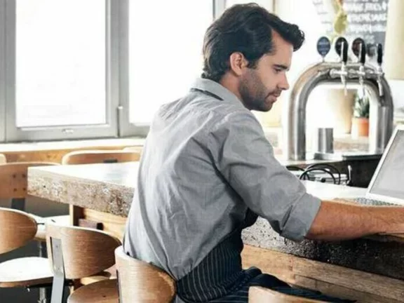 No balcão de um bar, onde há visivelmente uma chopeira e várias canecas de vidro, um homem de avental trabalha em um computador portátil (notebook) e anota com uma caneta em uma prancheta