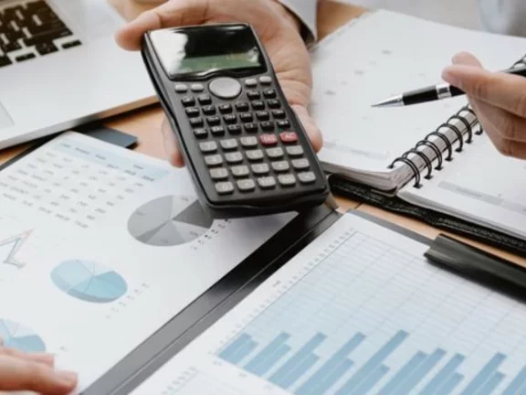 Profissionais utilizam cadernos, pranchetas com gráficos, um computador portátil (notebook) e uma calculadora para trabalhar