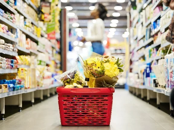 Uma cesta de compras no chão de um supermercado, enquanto uma mulher faz compras ao fundo
